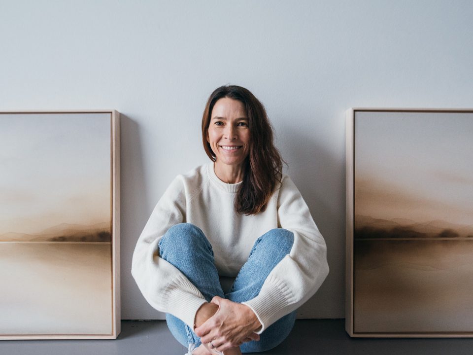 Carrie Loeb sitting cross legged with examples of her paintings leaned against the wall behind her